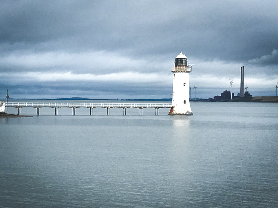 Phare blanc avec une longue jetée sur des eaux calmes et un arrière-plan industriel.