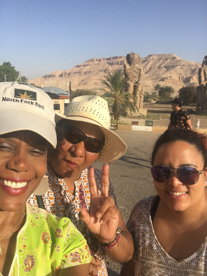 Trois femmes souriantes, posant à l'extérieur avec des ruines antiques en arrière-plan.