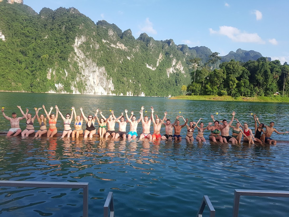 A group of people sitting on a dock with their hands up, with mountains and a lake in the background.