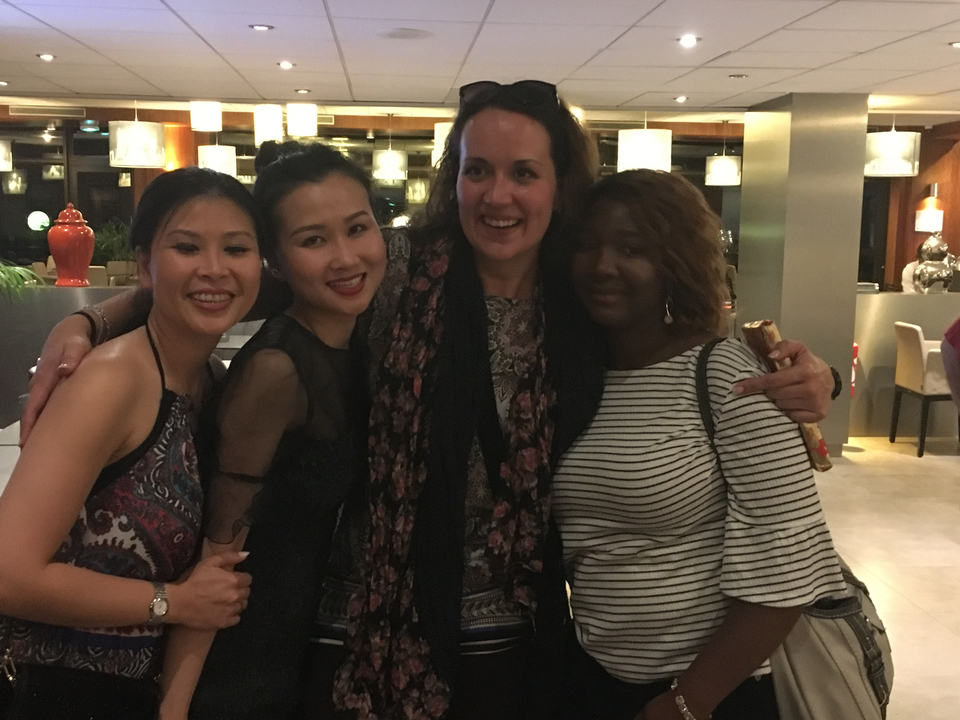 Quatre personnes debout bras dessus bras dessous à l'intérieur, avec des lampes suspendues au plafond.