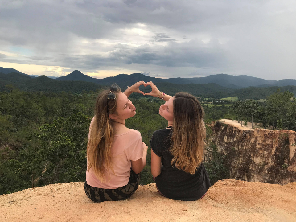 Two women making a heart shape with their hands against a scenic backdrop.