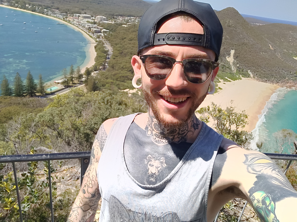 Man taking a selfie with a panoramic view of a beach and greenery.