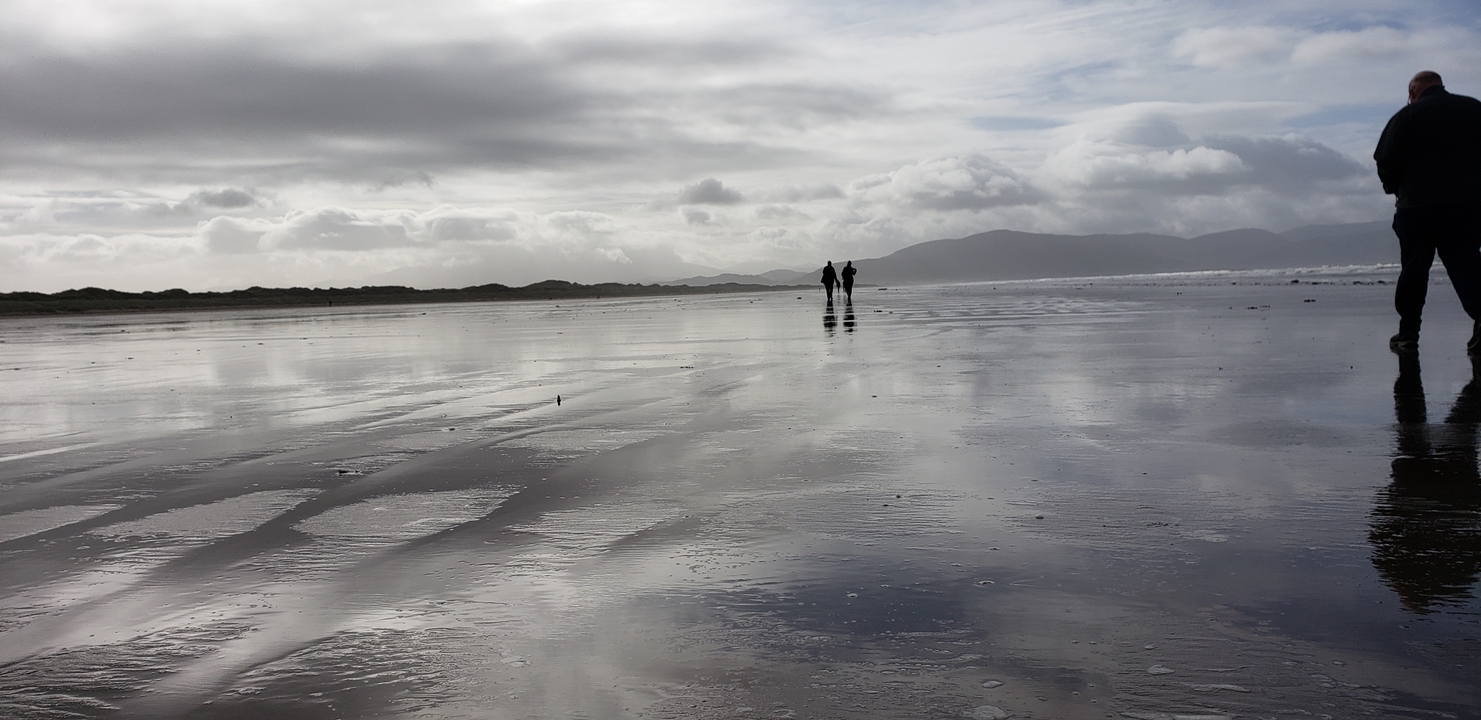 Large plage avec des reflets du ciel dans le sable mouillé, deux personnes marchant au loin.