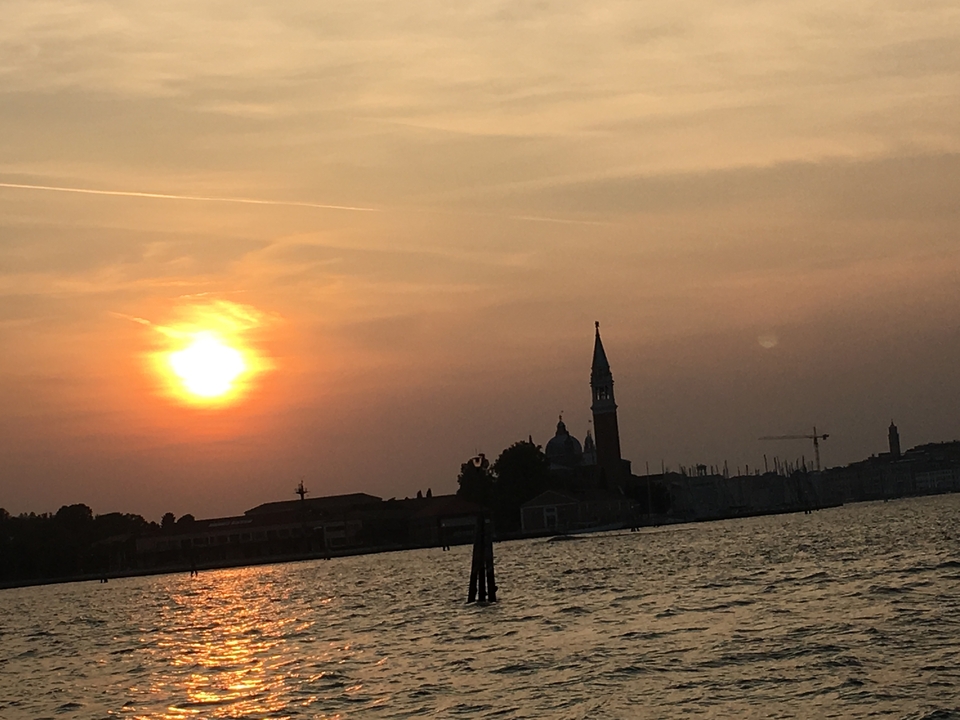 Vue du coucher de soleil sur une étendue d'eau avec des silhouettes de bâtiments, probablement à Venise.