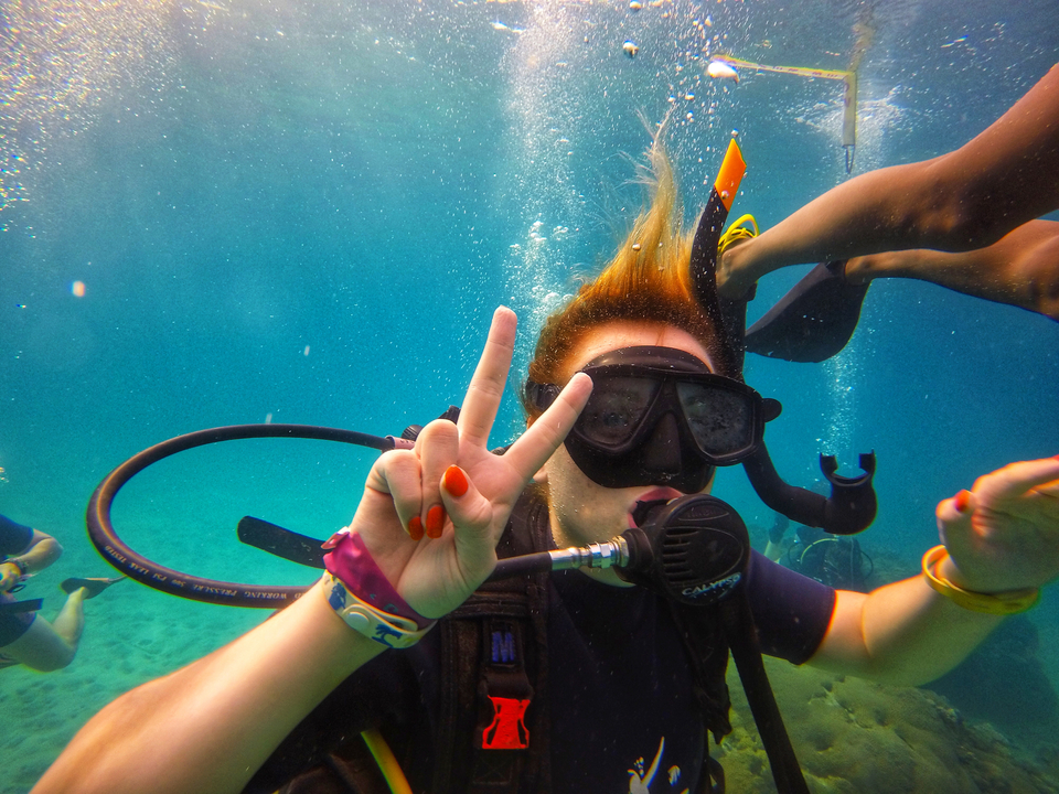 A diver underwater making a peace sign, with colorful bracelets visible.