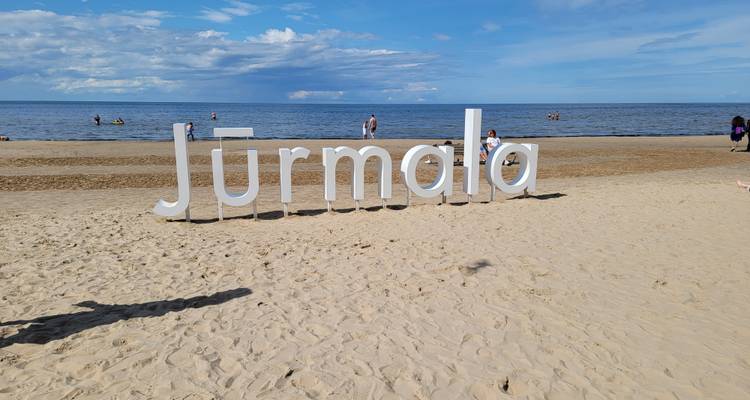 Strand mit 'Jurmala'-Schild, Menschen genießen den sonnigen Tag.
