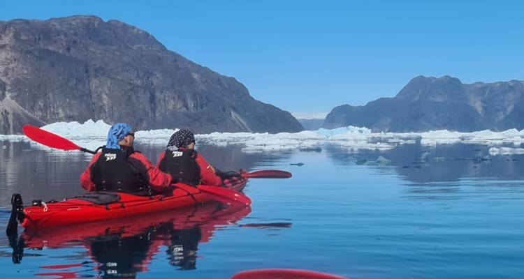 Two people kayaking among icebergs in a calm sea.