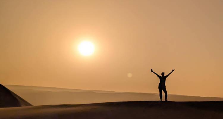 Een silhouet van een persoon die de armen omhoog steekt onder een ondergaande zon in een woestijn.