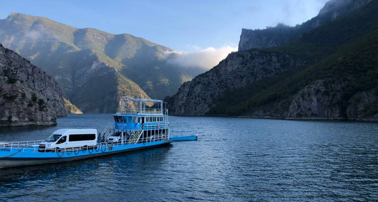 Blue ferry carrying a van on a scenic lake surrounded by mountains.