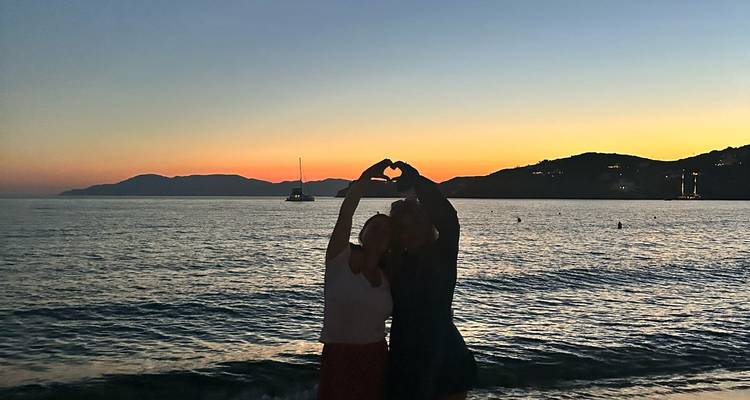 Deux personnes sur une plage au coucher du soleil formant un cœur avec leurs mains.