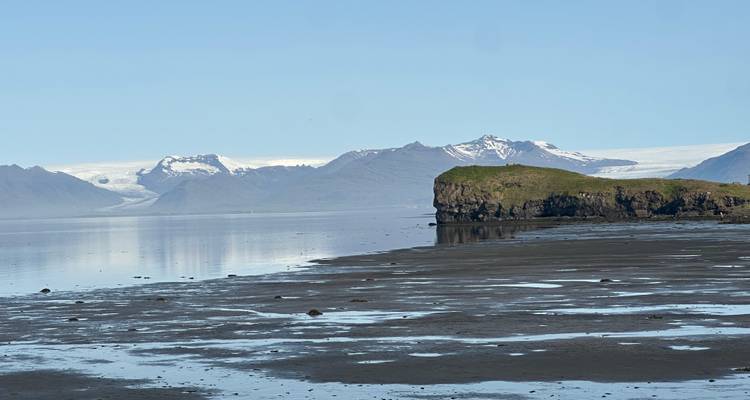 A scenic view of a coastal landscape with mountains and a grassy cliff reflected on calm water.