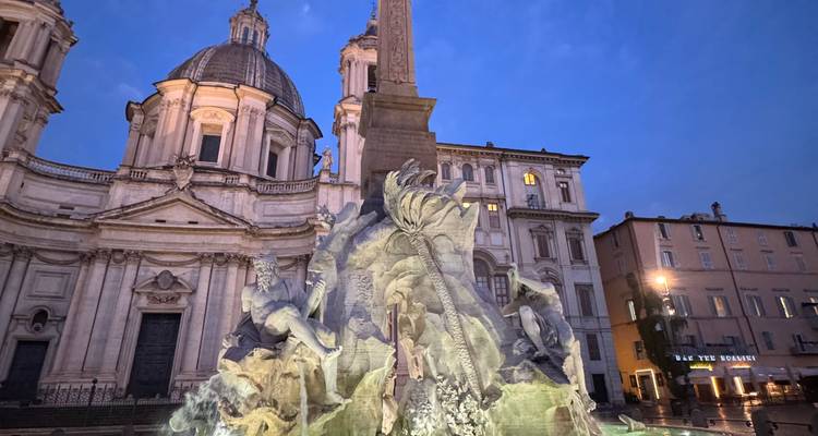 Une magnifique fontaine avec des statues et un obélisque sur une place illuminée en début de soirée.