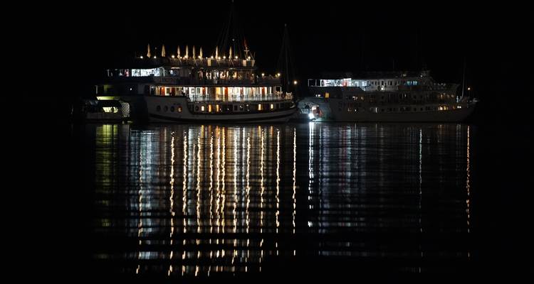Bateaux illuminés de lumières colorées la nuit, se reflétant sur l'eau.
