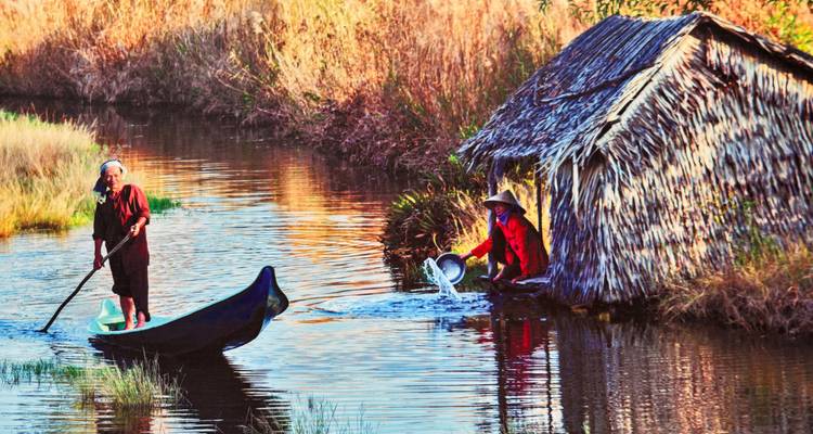 Zwei Menschen an einem Fluss, einer auf einem Boot und ein anderer bei einer strohgedeckten Hütte.