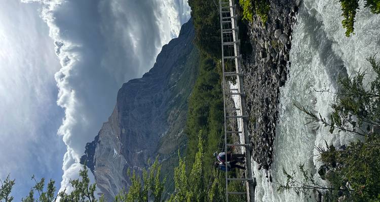Twee mensen die een brug over een rivier met bergen oversteken.