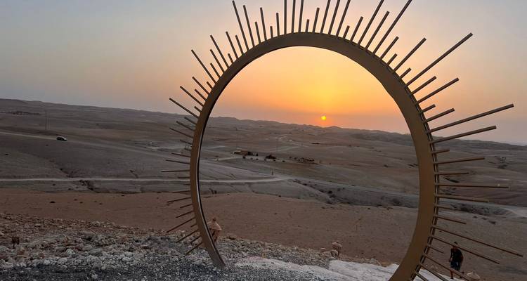 Woestijnlandschap met een grote metalen zonnesculptuur bij zonsondergang.