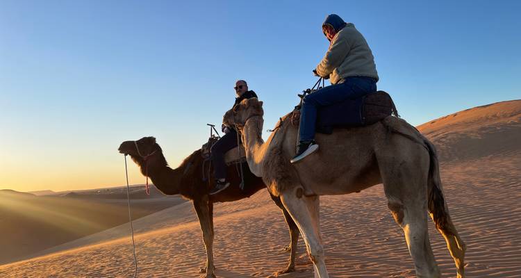 Zwei Menschen reiten auf Kamelen in der Wüste bei Sonnenuntergang.