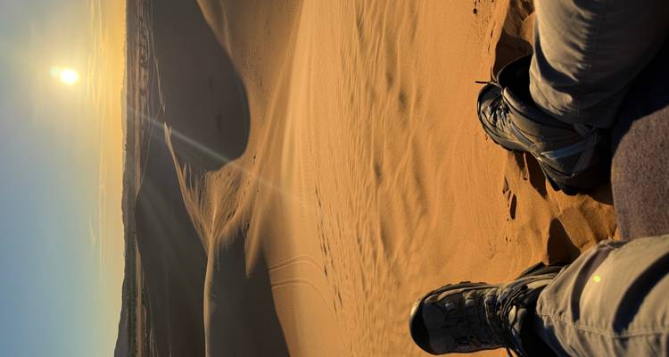Vista de dunas de arena durante el atardecer con una persona usando botas de senderismo en primer plano.