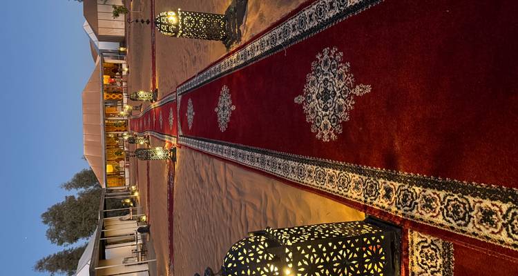 Sendero bordeado con elegantes faroles y alfombras rojas que lleva a una tienda en el desierto al atardecer.