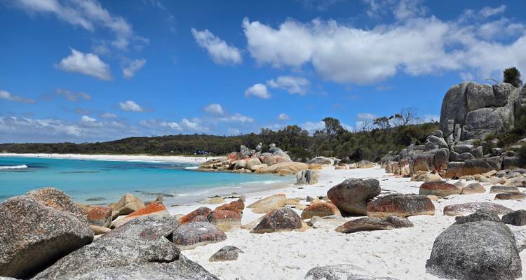 Felsen entlang eines weißen Sandstrandes mit klarem blauem Wasser unter einem sonnigen Himmel.