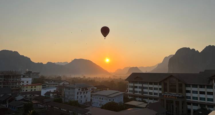 Globo aerostático silueteado flota sobre montañas kársticas brumosas al amanecer dorado sobre un pequeño pueblo.
