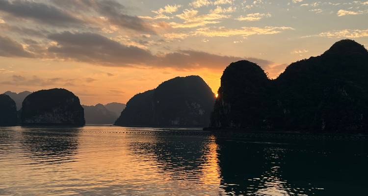 El atardecer anaranjado proyecta un resplandor cálido sobre las aguas tranquilas y las imponentes islas de piedra caliza de la Bahía de Ha Long.