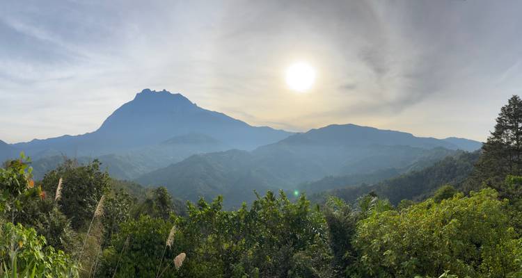 Panoramic morning view of Mount Kinabalu rising over rolling green hills with the sun shining above.