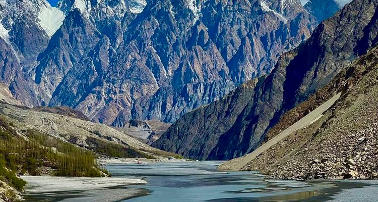Dramatic snow-capped Karakoram peaks tower over a winding river in a steep rocky gorge.