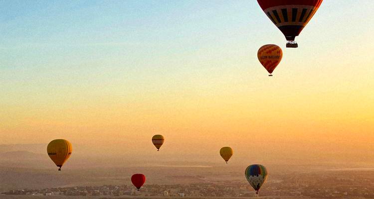 Multiple colourful hot-air balloons floating above a hazy desert cityscape at sunrise.