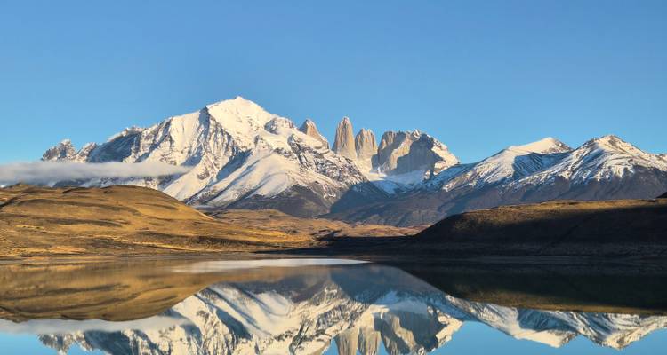 Snowy Torres del Paine massif perfectly reflected in a still lake at dawn, with golden foothills in the foreground.