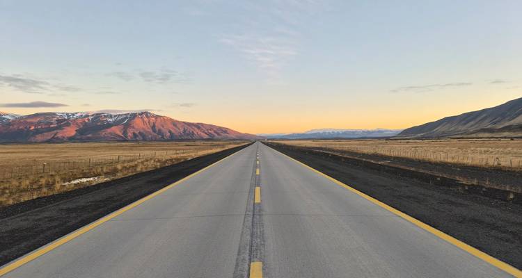 A long straight highway disappears into a pastel sunrise horizon between rugged Patagonian ranges.