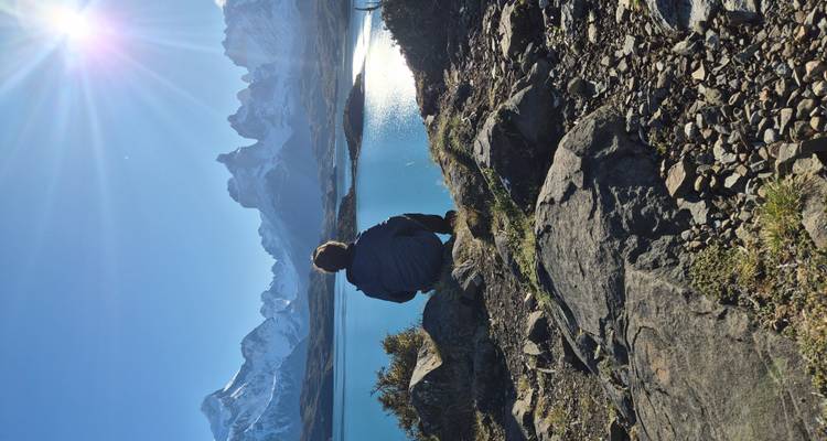A hiker sits on a rocky outcrop admiring bright blue Patagonian lake waters with towering snowy peaks beyond.