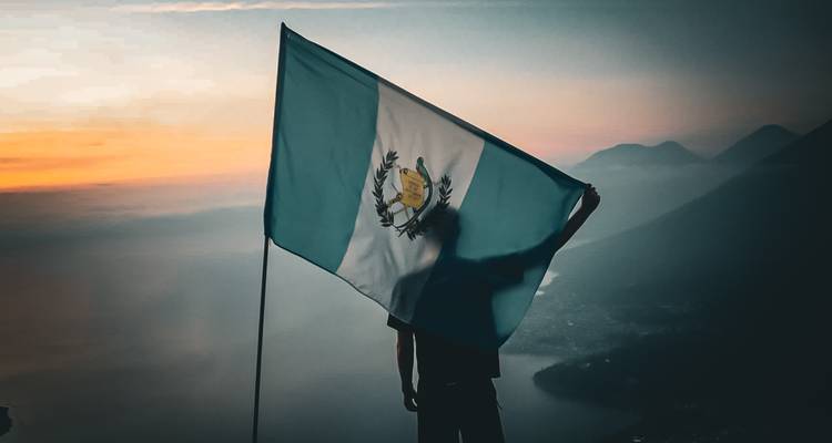 Silhouet van een wandelaar die de Guatemalaanse vlag omhoog houdt op een bergkam bij kleurrijke dageraad.