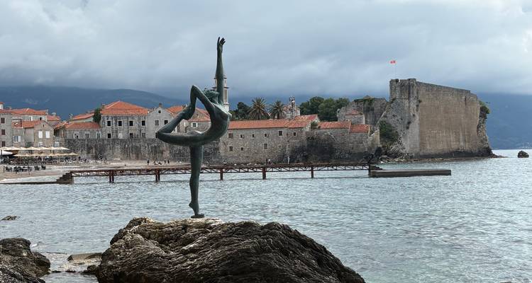 Estatua de una bailarina elegante en una roca con las murallas y torres de la Ciudad Vieja de Budva al otro lado de una bahía tranquila.