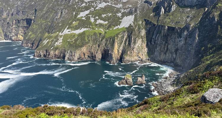 Dramatic high sea cliffs covered with green patches dropping into swirling blue Atlantic waters.