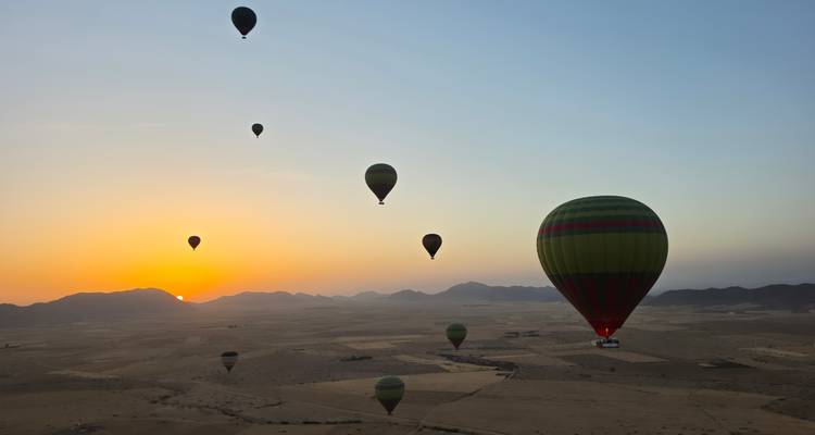 Múltiples globos aerostáticos de colores brillantes flotan sobre una meseta árida con un amanecer resplandeciente sobre las montañas lejanas.