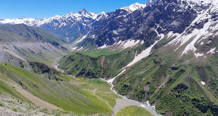 Valle alpino amplio con laderas verde exuberante, picos nevados y parches de nieve de final de temporada bajo un cielo azul despejado.