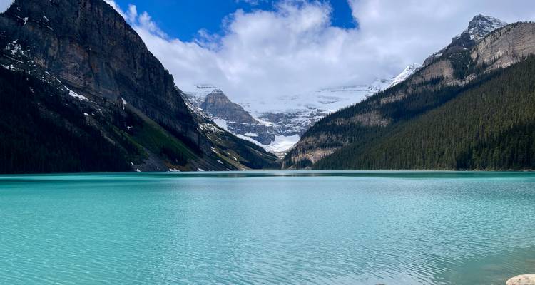 Der kristallklare Lake Louise erstreckt sich in Richtung aufragender Klippen und entfernter Gletscher unter einem fleckigen blauen Himmel.
