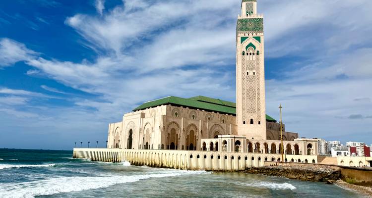 The Hassan II Mosque rises over crashing Atlantic waves beneath blue skies.
