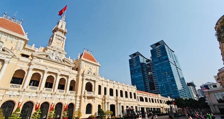 L'Hôtel de Ville de Ho Chi Minh City datant de l'époque coloniale avec le drapeau vietnamien se détachant sur un ciel bleu dégagé, mis en contraste avec une tour de verre moderne.