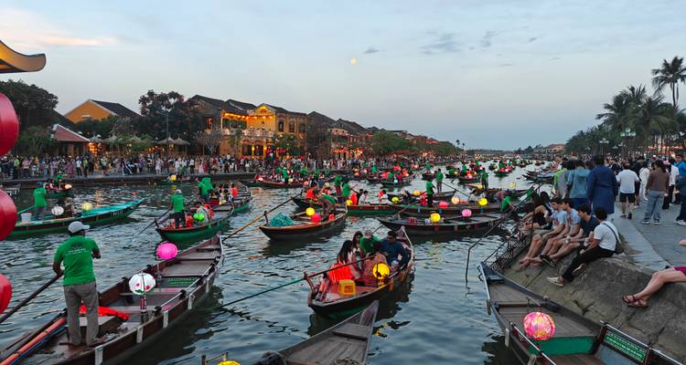 Scène en soirée à Hoi An avec des dizaines de bateaux illuminés par des lanternes sur le fleuve et des foules le long de la promenade sous une lune montante.