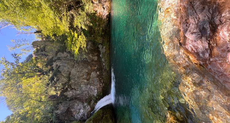 Klare türkisfarbene Wasserbecken unter einem kleinen kaskadierenden Wasserfall, umgeben von üppigem grünem Wald und Felswänden.