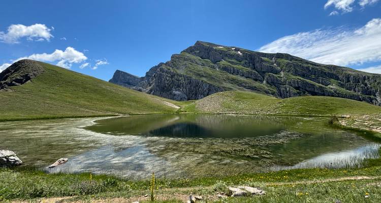 A crystal-clear alpine lake mirrors jagged green mountains under a bright blue sky.