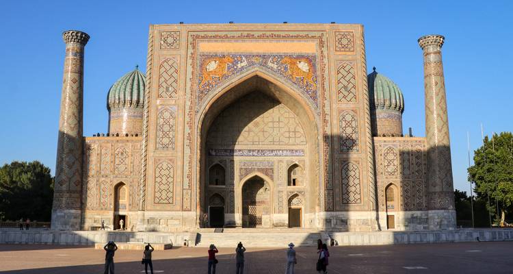 Majestic Sher-Dor Madrasa at Registan Square with visitors photographing the facade in evening light.