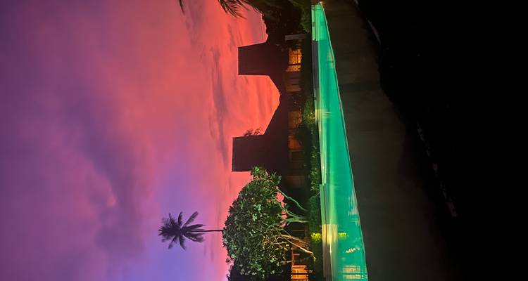 Infinity pool glowing green against vibrant pink and purple sunset skies with tropical palm and wooden villas