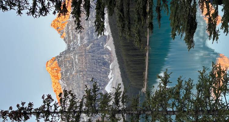 Les pics montagneux baignés de soleil se reflétant dans le lac turquoise à travers les arbres en silhouette au lever du soleil