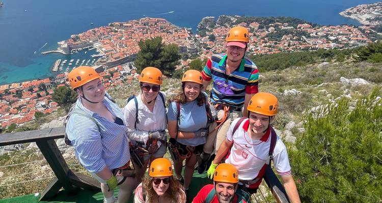 Thrilled travellers in helmets stand on a mountaintop zipline platform with aerial view of Dubrovnik’s old town and sea below.