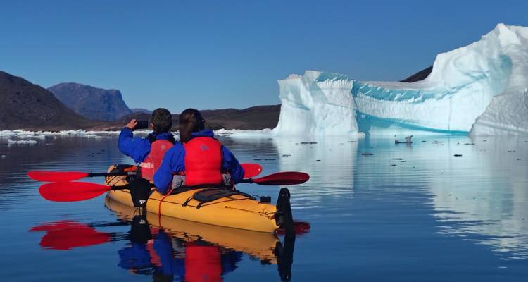 Dos kayakistas reman pasando un iceberg imponente reflejado en las tranquilas aguas del Ártico.