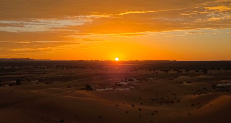 Le soleil d'un orange éclatant se couche sur les dunes de sable ondulantes du Sahara, avec de minuscules tentes de campement désertique visibles au loin.