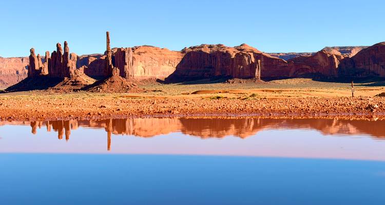 Red sandstone buttes of Monument Valley reflected in a still rain-fed pool under a clear blue sky.